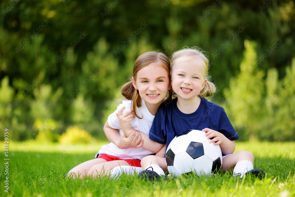 Two cute little sisters having fun playing a soccer game on sunny summer day. Sport activities for children. Two cute little sisters having fun playing a soccer game on sunny summer day. Sport activities for children.