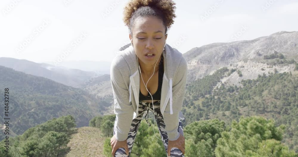 Young attractive woman in sportswear with headphones bending on knees taking rest with beautiful mountains on background. 