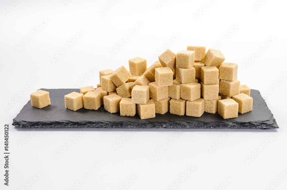 Pile of cane sucar cubes and one cube alone on rectangular dark shale plate