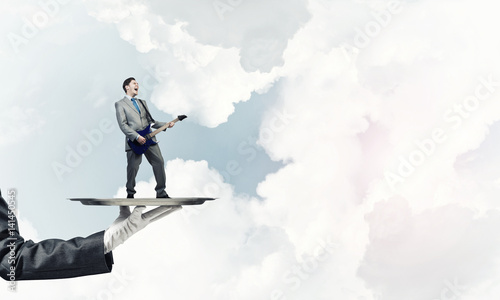 Businessman on metal tray playing electric guitar against blue sky background © adam121