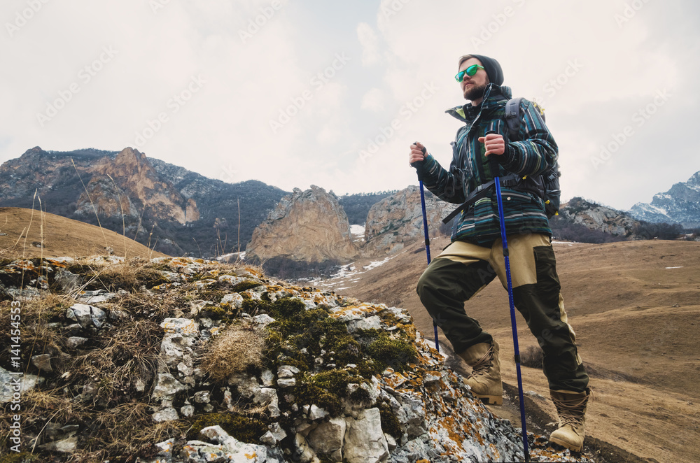 Obraz premium Tired bearded hipster with binoculars in his hands sits on a stone among the mountains and looks out into the distance