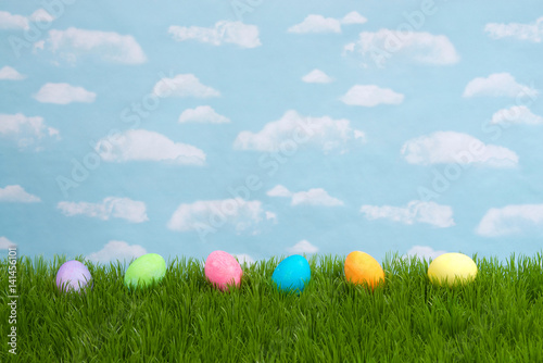 Colorful Easter Eggs lined up in a row on tall grass with blue background sky with clouds. Copy space