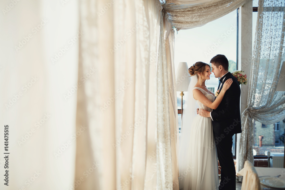 Groom holds bride's waist tender standing with her before panoramic window Groom holds bride's waist tender standing with her before panoramic window