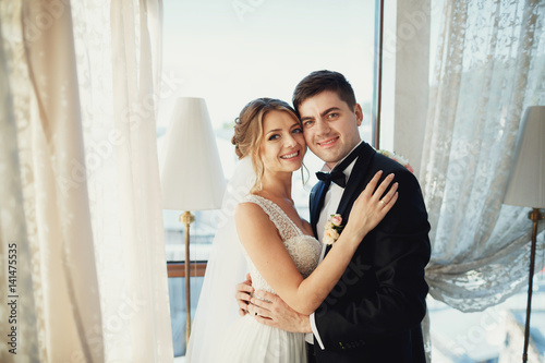 Handsome brunette groom and dark blonde bride hug before bright window © myronovychoksana