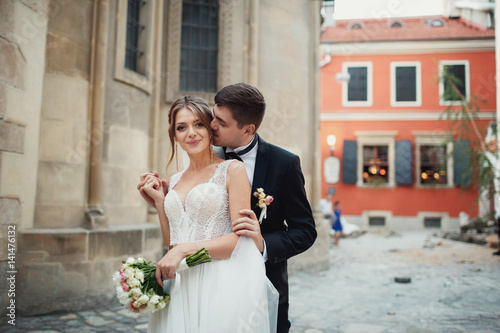 Groom kisses pretty bride from behind standing with her on the frontyard of an old church