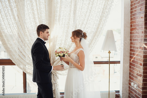Groom gives wedding bouquet to the bride standing before panoramic window