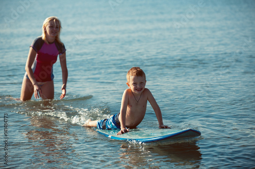 Young woman teaching surfing her son in the ocean in a sunny day © Kirill Grekov