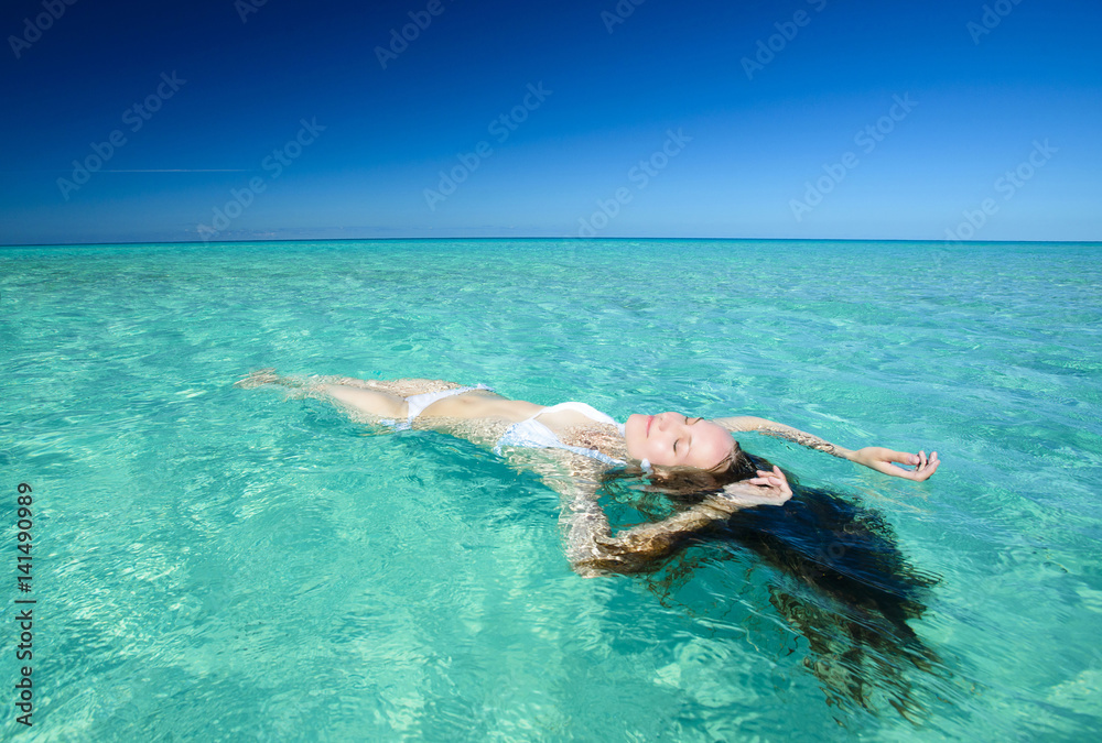 Beautiful young brunette woman with long hair dressed in white swimsuit is lying on the crystal azure water of caribbean sea