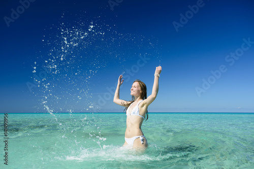 Beautiful young brunette woman with long hair dressed in white swimsuit is splashing on the crystal azure water of caribbean sea