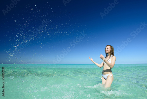 Beautiful young brunette woman with long hair dressed in white swimsuit is standing and smiling on the crystal azure water of caribbean sea