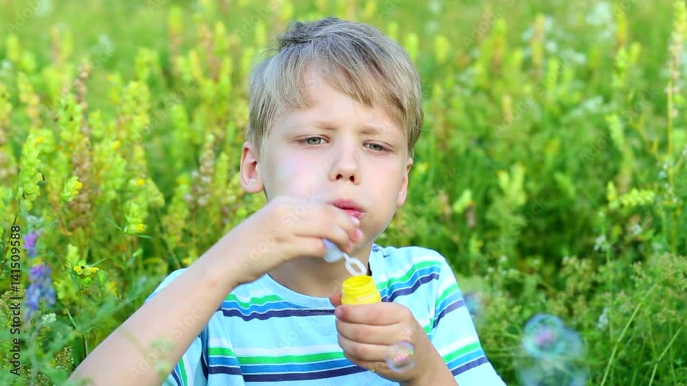 Closeup portrait of funny cute little kid playing outside in beautiful summer meadow. Child blows soap bubbles cheerfully while sitting in beautiful green grass. Real time full hd video footage. Closeup portrait of funny cute little kid playing outside in beautiful summer meadow. Child blows soap bubbles cheerfully while sitting in beautiful green grass. Real time full hd video footage.