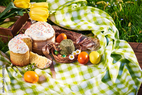 Easter cake and bright colored eggs gathered together with meat, treats and tulips on the lawn among the grass