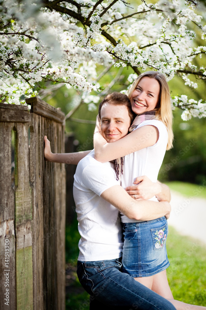 Fototapeta premium Tender couple in love smell blooming cherry tree with white flowers in the park in spring. Happy girl and guy smile and hug outdoor, they are wearing in jeans, white t-shirts.