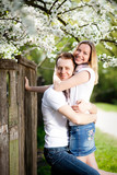 Tender couple in love smell blooming cherry tree with white flowers in the park in spring. Happy girl and guy smile and hug outdoor, they are wearing in jeans, white t-shirts.