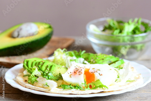 Healthy breakfast idea. Delicious tortilla with a poached egg, avocado slices, napa cabbage, salad mix, sauce and spices on a plate. Avocado half, green salad in a glass bowl on a wood table. Closeup