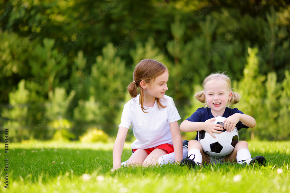 Two cute little sisters having fun playing a soccer game on sunny summer day. Sport activities for children. Two cute little sisters having fun playing a soccer game on sunny summer day. Sport activities for children.