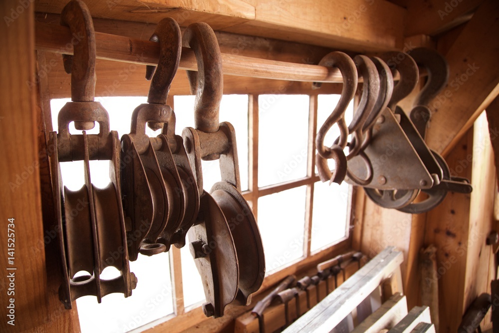 Hanging rusted heavy cast iron hooks attached to pulley wheel on wood rod  by window light in traditional rustic industrial woodworking workshop  Photos