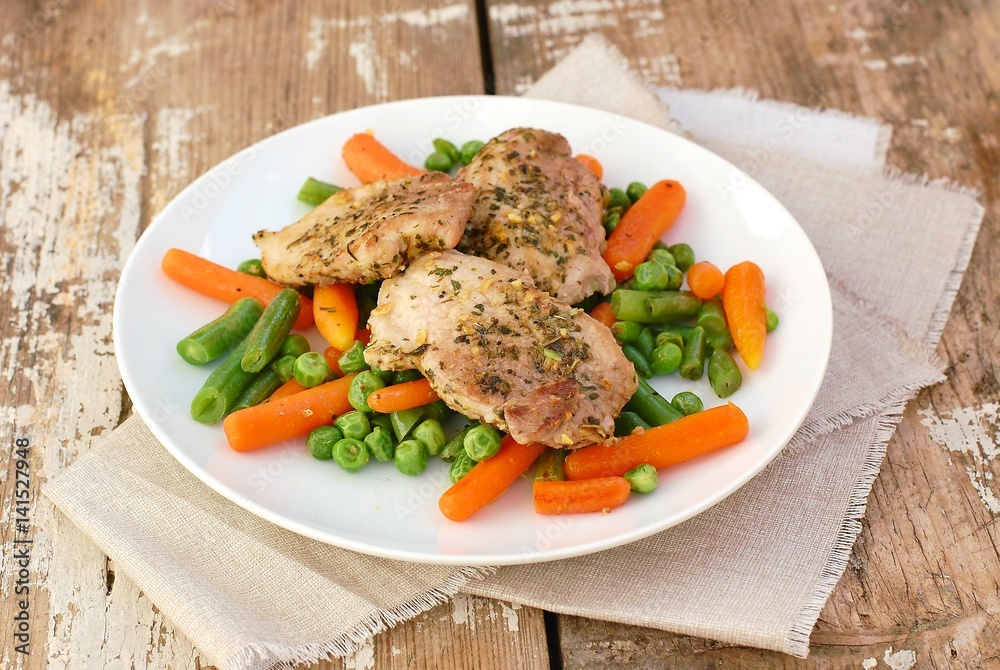 Food from pork meat fillet with spices and spring vegetable baby carrots, peas and green beans on white plate on wooden background