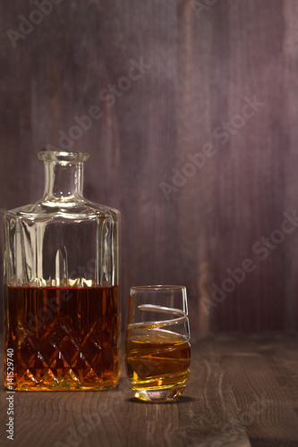 A glass of short with whiskey stands on a wooden table near a decanter