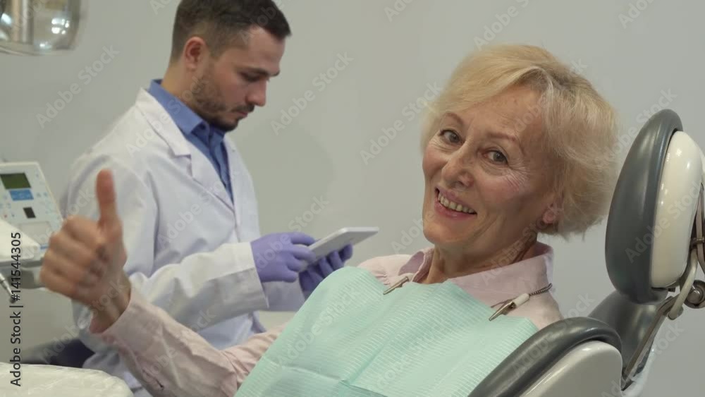 Senior female client showing her thumb up on the dental chair. Attractive caucasian woman approving apppointment at the dentist. Camera tilting up along aged blond lady smiling for the camera against Senior female client showing her thumb up on the dental chair. Attractive caucasian woman approving apppointment at the dentist. Camera tilting up along aged blond lady smiling for the camera against