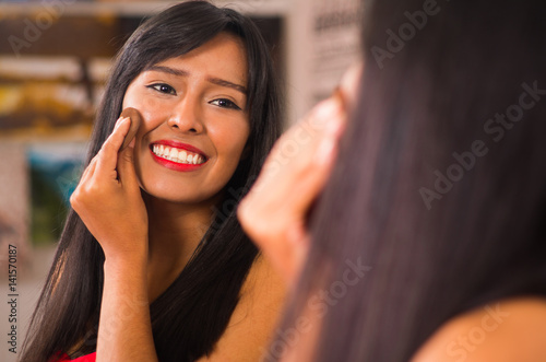 Beautiful brunette applying make up while smiling happily, seen from behind and face reflecting in mirror
