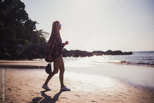 Young beautiful girl in stylish sunglasses and with a fashionable bag at sunset walking on a beach