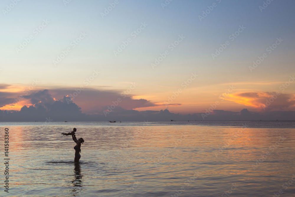 Silhouette of mother with her baby against the sunset and lens flare at sea. Asian family activity lifestyle.