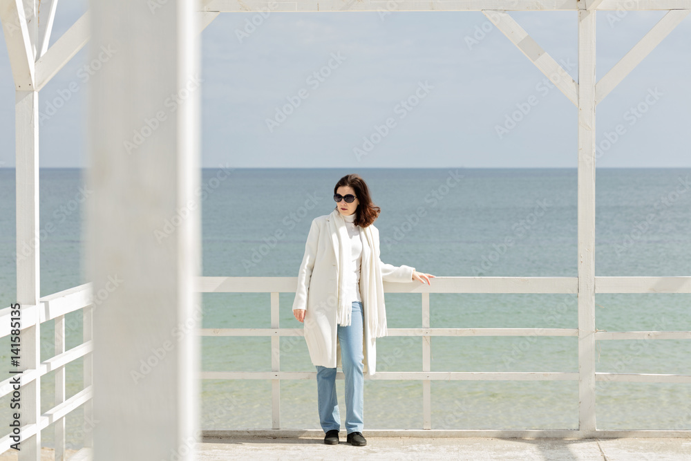 Young woman (brunette) in a white coat on the promenade near the sea.