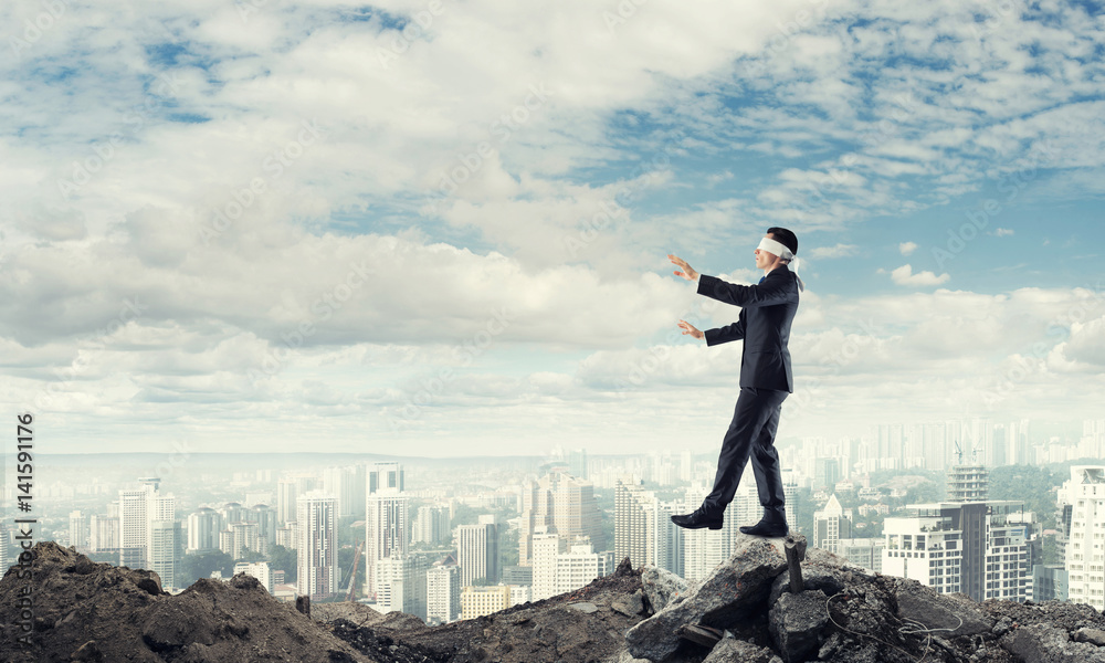 Young businessman in blindfold walking carefully and cityscape at background Young businessman in blindfold walking carefully and cityscape at background
