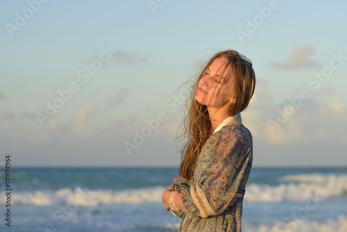 Beautiful young blond woman in dress stands on the beach of caribbean sea and watches at sunset
