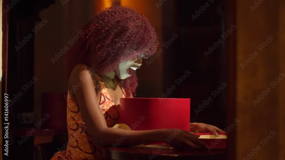 Horizontal close-up portrait of surprised afro-american girl with red curly hair by the present in the round red box with golden ribbons, sitting near the window in the cosy cafe