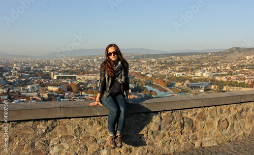 Traveler girl wearing black leather jacket sitting on the stone wall. Panoramic view of old Tbilisi, Georgia at the background. The view from the castle Narikala.