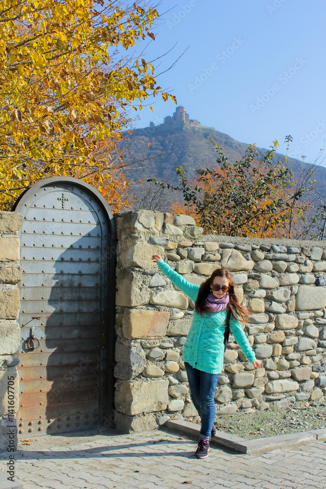 Fototapeta premium Traveler girl with hands up walking near old stone wall. Jvari Monastery on the hill at the background. Woman traveler walking in city Mtskheta, Georgia
