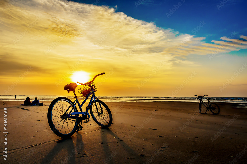Fototapeta premium Silhouette of bikes on beach at sunrise at an ocean with people in the distance