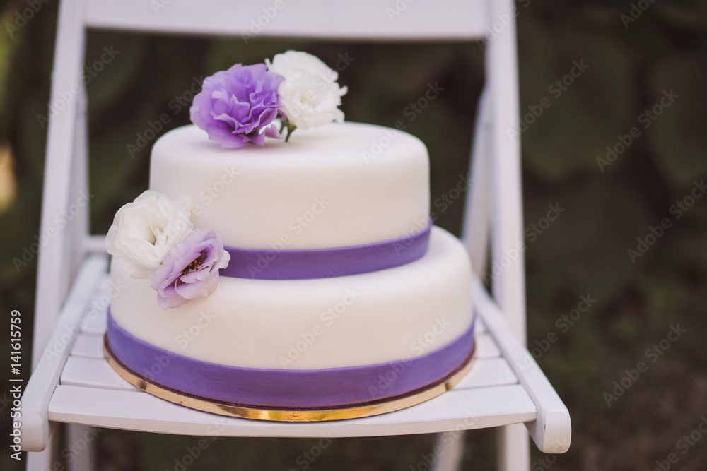 White wedding cake with purple flowers decoration outdoors on wooden modern chair. Front view, copy space. White wedding cake with purple flowers decoration outdoors on wooden modern chair. Front view, copy space.