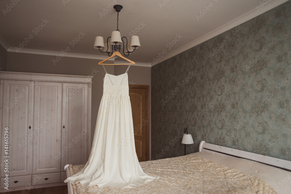 Modeern bridal fashion. White light wedding dress hanging in bedroom. Modeern bridal fashion. White light wedding dress hanging in bedroom.
