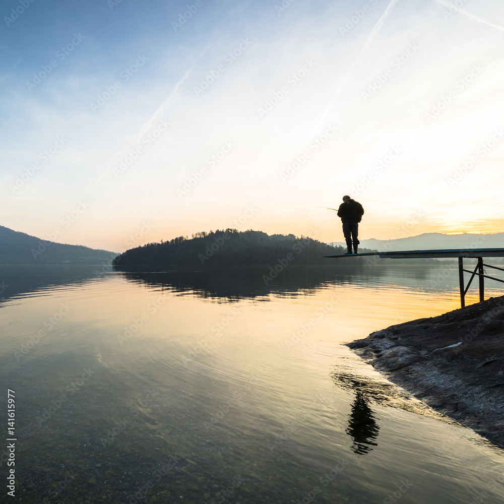 A man catches a fish. Happy relaxed hours alone with nature. Landscape of a mountain lake. Evening light of the setting sun. Male silhouette with a fishing rod in the backlight.