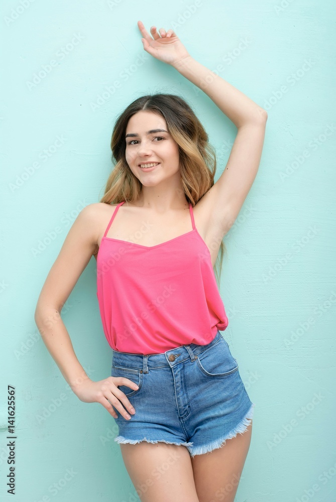 Young girl with long curly hair posing on blue background in studio. She wears shorts, pink T-shirt, pink sunglasses. She keeps hat overhead, holds blue skateboard on booty, looks excited. Young girl with long curly hair posing on blue background in studio. She wears shorts, pink T-shirt, pink sunglasses. She keeps hat overhead, holds blue skateboard on booty, looks excited.