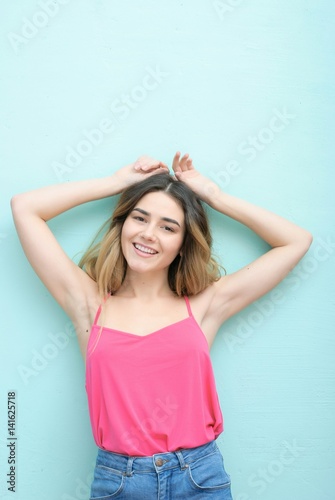 Young girl with long curly hair posing on blue background in studio. She wears shorts, pink T-shirt, pink sunglasses. She keeps hat overhead, holds blue skateboard on booty, looks excited.