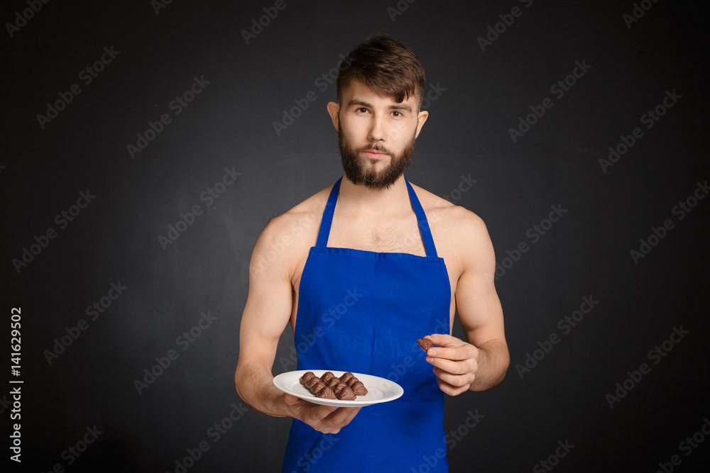 Hot sexy naked handsome man with chocolates, chocolate candies. Smiling naked handsome man wearing dark blue apron holding a white plate with many sweets, chocolate candies. Handsome confectioner. Hot sexy naked handsome man with chocolates, chocolate candies. Smiling naked handsome man wearing dark blue apron holding a white plate with many sweets, chocolate candies. Handsome confectioner.