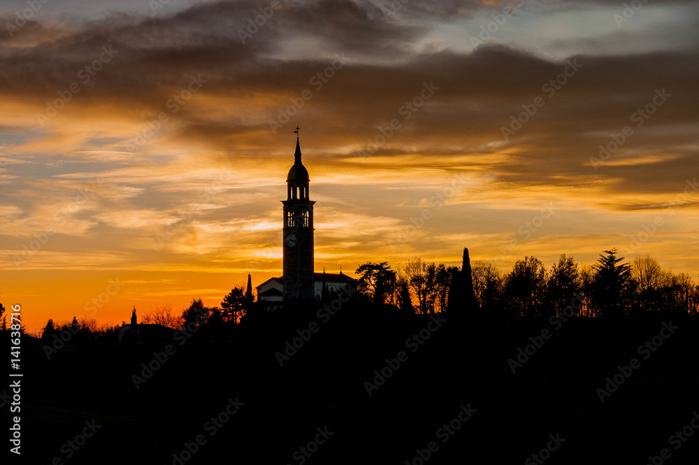 Evening sunset with black silhouette of small church and bell tower and trees on colorful flaming cloudy sky background.