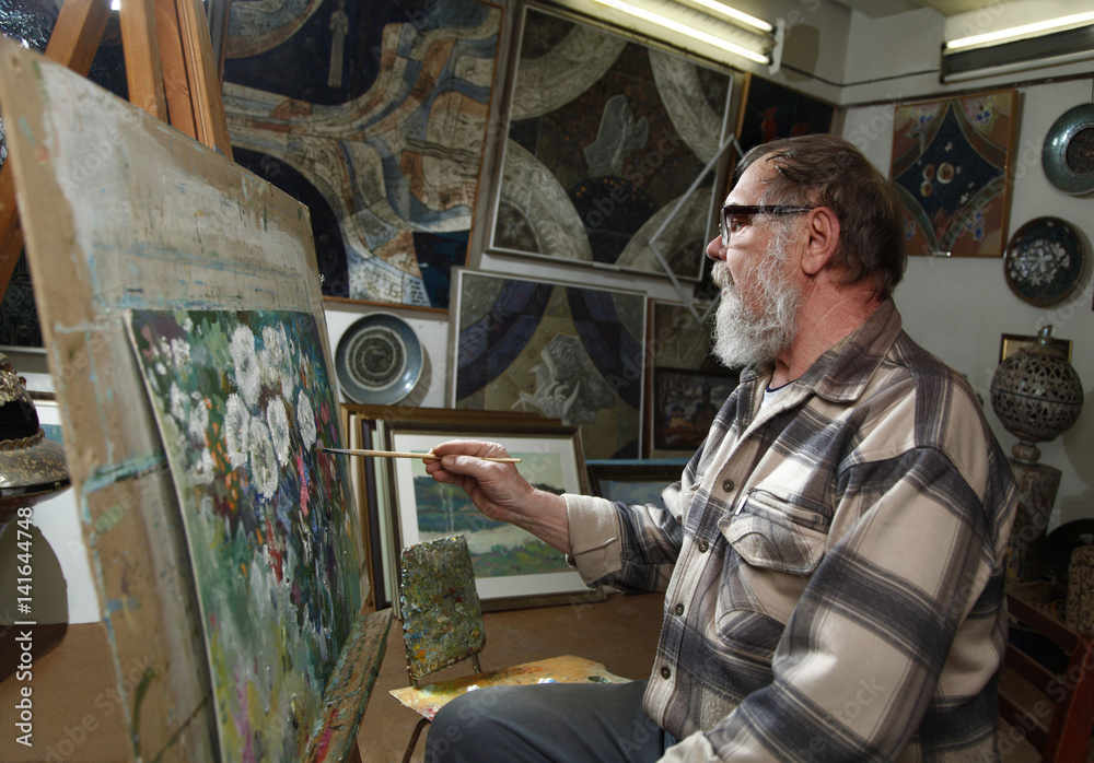 Elderly painter with beard and glasses draws a flowers picture by oil paint in art workshop Elderly painter with beard and glasses draws a flowers picture by oil paint in art workshop