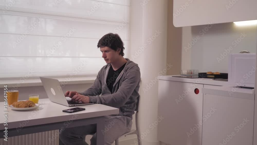 Man sitting at the desk with cup and croissant on white place. Male working looking on screen computer in kitchen. Businessman computing at home in the morning. Man sitting at the desk with cup and croissant on white place. Male working looking on screen computer in kitchen. Businessman computing at home in the morning.