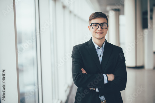 Confident businessman standing with crossed hands against office windows