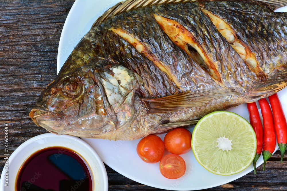 Grilled fish on white plate with chili pepper, lemon and tomato on wooden background, top view. Grilled fish on white plate with chili pepper, lemon and tomato on wooden background, top view.