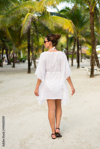 Back view on woman on tropical vacation at caribbean resort beach. Brunette model wearing summer fashion sunglasses and white kaftan. Riviera Maya  Mexico.