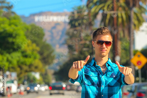 Cool guy standing on the boulevard showing thumbs up with a Hollywood sign behind him. Beautiful perfect life. photo