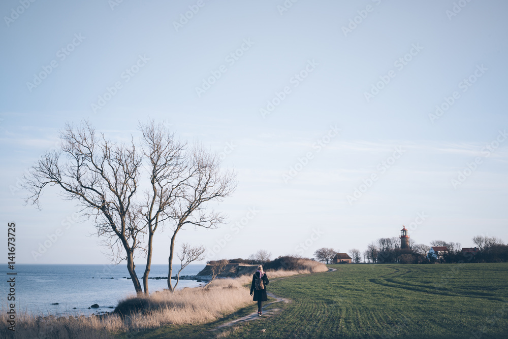 woman with backpack walking along path on high coast cliff with sea and lighthouse in background woman with backpack walking along path on high coast cliff with sea and lighthouse in background