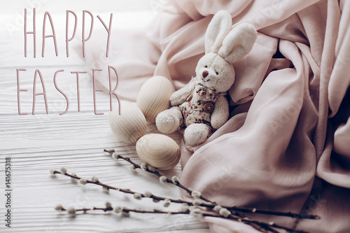 Fototapeta Naklejka Na Ścianę i Meble -  happy easter text greeting card sign on stylish easter bunny rabbit and eggs and willow buds on rustic wooden background and fabric . with space for text, top view. soft light