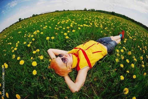 Art portrait with the fisheye effect of a young pretty blonde woman with short hair in yellow-red vest, wearing sunglasses, relaxing on meadow of yellow flowers of dandelions. photo